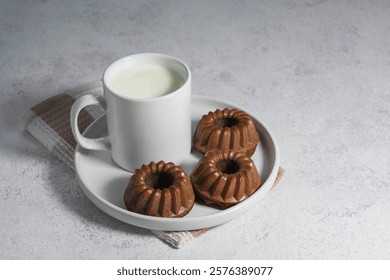 A rustic food composition featuring three chocolate mini bundt cakes served with milk on a white plate. Styled on a light textured background with a cozy feel.