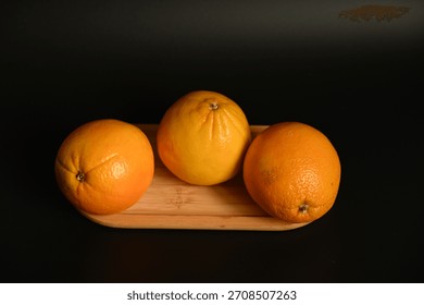 Rustic food presentation featuring three oranges on a small wood board. The dark background emphasizes the bold, vibrant color of the fruit, suitable for cooking, juicing, and raw food themes.