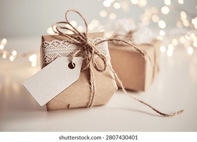 Rustic gift box wrapped in kraft paper with twine and lace ribbon

Two beautifully wrapped holiday gifts on a bright surface with warm, glowing bokeh lights in the background. The foreground present features brown kraft wrapping paper, an elegant white lace trim, a blank paper gift tag, and a natural jute twine bow. Perfect for Christmas, wedding favors, Valentine's Day, anniversaries, and cozy winter celebrations.