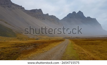 Rustic gravel path leads through vast, golden field towards historic replica of Viking village, nestled dramatically at base of the misty, imposing Vestrahorn mountain range in southeastern Iceland