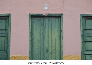 A rustic green wooden door with weathered paint stands at the center of a pastel pink facade. Flanked by two matching green wooden windows, the scene features an old, vintage architectural style - Powered by Shutterstock