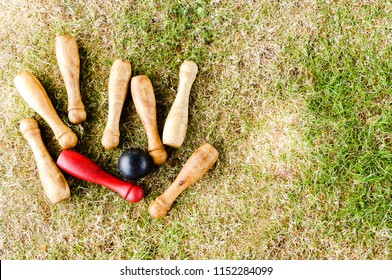 Rustic hand made set of discarded outdoor skittles laying on sun baked grass. One red pin, with seven other natural coloured pins, black stained bowling ball. Garden game, England.