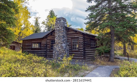 A rustic historic log cabin featuring a tall river-stone chimney stands nestled among autumn trees in Grand Teton National Park, Wyoming. The dark hand-hewn log walls contrast beautifully with the golden yellows and greens of surrounding aspen and conifer trees. Dramatic cloud-streaked skies frame the scene, evoking the rugged pioneer heritage of Jackson Hole.