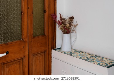 Rustic home decor featuring a white pitcher with dried flowers on a colorful mosaic tile surface next to a wooden door.