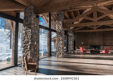 Rustic interior of an alpine hotel featuring wooden beams, stone pillars, a modern fireplace, red armchairs, and snowy trees visible through large windows.
