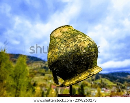 The rustic jug stands like an ancient relic against the backdrop of high mountains, under a blue sky with clouds. A nostalgic combination of nature and history in a picturesque composition.