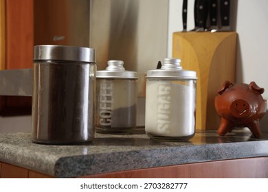 Rustic kitchen countertop with coffee jars and sugar canister, brown metal tin, vintage ceramic piggybank beside wooden knife block, soft warm lighting