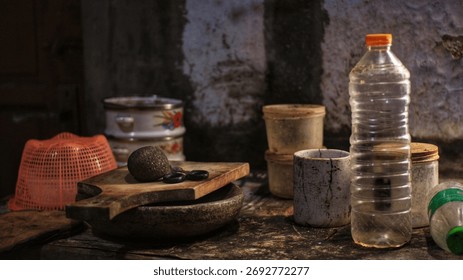 Rustic kitchen table with cutting board, stone mortar, bowls, and a plastic bottle in moody light against an aged wall. Traditional home interior still life.