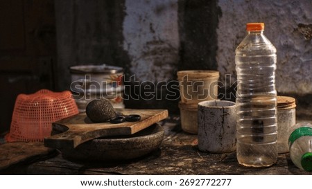 Rustic kitchen table with cutting board, stone mortar, bowls, and a plastic bottle in moody light against an aged wall. Traditional home interior still life.