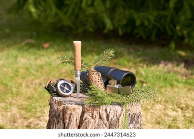 A rustic knife with a wooden handle, a vintage monocular, a lighter, a handheld compass, and a pine branch with a cone rest on a wooden stump against a blurred background of lush green grass. - Powered by Shutterstock