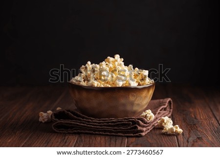 Rustic lifestyle composition featuring a brown ceramic bowl filled with freshly popped popcorn, placed on a folded cloth atop a wooden table. 