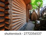 A rustic log cabin with a cobblestone path leads to a small wooden shed. A rock fountain sits beside a wrought iron fence, shaded by lush green trees.