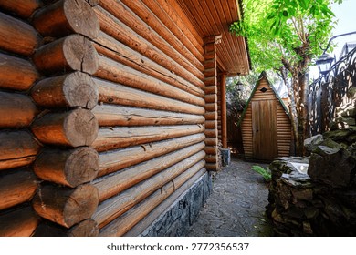 A rustic log cabin with a cobblestone path leads to a small wooden shed. A rock fountain sits beside a wrought iron fence, shaded by lush green trees.