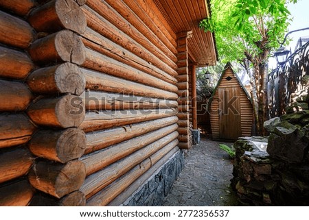 A rustic log cabin with a cobblestone path leads to a small wooden shed. A rock fountain sits beside a wrought iron fence, shaded by lush green trees.