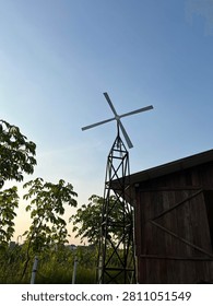 A rustic metal windmill tower standing beside a wooden barn in a rural landscape under a clear blue sky at golden hour. Surrounded by green trees and farmland, the scene conveys peaceful countryside living, sustainable energy, agriculture, and nature with ample copy space.