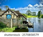 Rustic millhouse situated by a tranquil lake, blending natural beauty with old-world craftsmanship. The millhouse is constructed with weathered stone walls, a thatched roof, and a striking blue door.