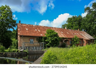 Rustic old mill building with a rusty, corrugated metal roof, set beside a tranquil waterway. The structure is made of weathered brick and stone, partially covered with green vegetation. Large trees surround the building, contributing to a lush, natural setting. A small dam or sluice gate is visible along the waterway, indicating the mill’s historical function in water management or grain processing. The scene evokes a peaceful, rural atmosphere under a bright blue sky with scattered clouds.