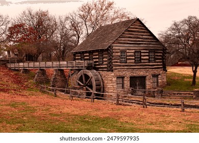 A rustic old mill with a large water wheel sits serenely amidst autumn colors. Stone and wood construction give it a charming, historical look, reminiscent of bygone days.