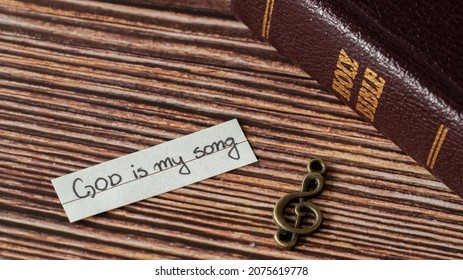 A rustic old treble clef note, handwritten quote: God is my song, and closed Holy Bible on wooden table. Christian biblical concept of music, praise, worship, and joy in the LORD. A closeup.