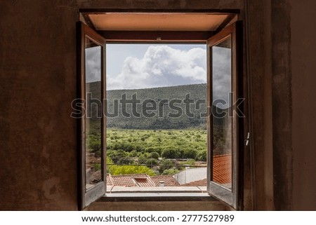 Rustic open window framing a scenic landscape of green hills and countryside in Mira de Aire, Portugal, with dramatic clouds and traditional village rooftops.