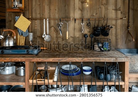 Rustic outdoor camp kitchen in a forest cabin with a wooden worktop, gas burner, steel kettle and utensils hanging from a brass rail. Enamel plates, pots and mugs are neatly arranged on open shelves, evoking off-grid glamping and slow countryside living