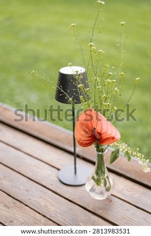 Rustic outdoor dining table arrangement with a glass vase of fresh wildflowers, black cordless table lamp, wooden salt and pepper grinders, and white napkin holder against.