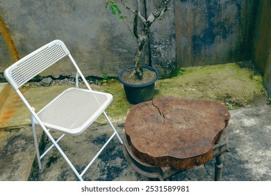 Rustic outdoor setting with a white folding chair, wooden table made from a tree stump, and potted plant against a weathered concrete wall. - Powered by Shutterstock