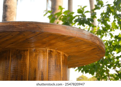 A rustic outdoor table made from a repurposed old wire spool, showcasing sustainable and creative furniture design. The table sits on a wooden deck, surrounded by natural greenery.