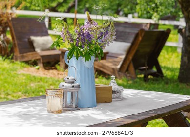 Rustic outdoor table setting decorated with bouquet of wildflowers in blue metal pitcher in garden patio relaxation area. Wooden furniture, relaxing countryside atmosphere, leisure, summer recreation.