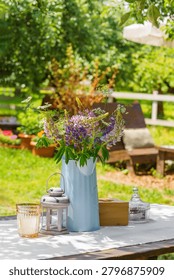 Rustic outdoor table setting decorated with bouquet of wildflowers in blue metal pitcher in garden patio relaxation area. Wooden furniture, relaxing countryside atmosphere, leisure, summer recreation.