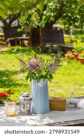 Rustic outdoor table setting decorated with bouquet of wildflowers in blue metal pitcher in garden patio relaxation area. Wooden furniture, relaxing countryside atmosphere, leisure, summer recreation.