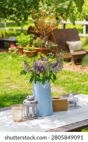 Rustic outdoor table setting decorated with bouquet of wildflowers in blue metal pitcher in garden patio relaxation area. Wooden furniture, relaxing countryside atmosphere, leisure, summer recreation.