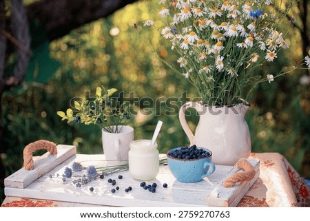 A rustic outdoor table setting features a bouquet of wildflowers, a jar of yogurt, and a bowl of ripe blueberries, evoking a sense of natural abundance and tranquility