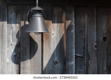 Rustic outdoor wall lamp casting shadow on weathered wooden fence in natural sunlight.