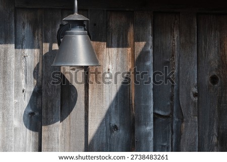 Rustic outdoor wall lamp casting shadow on weathered wooden fence in natural sunlight.