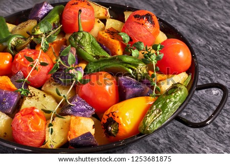 Rustic oven baked vegetables in black baking dish con gray stone background. Seasonal vegetarian vegan meal. Ingredients: potatoes, purple and sweet potatoes, tomatoes and peppers