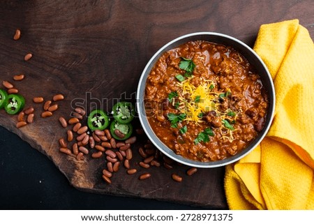 A rustic overhead view of homemade beef chili served in bowls and topped with shredded cheddar cheese, fresh herbs, and sliced jalapeños. Surrounded by red beans, chili flakes, and cornbread on a wooden table, this image conveys comfort food, hearty meals, and home cooking. Perfect for food blogs, recipe websites, cookbooks, restaurant menus, seasonal promotions, and advertising focused on warm, satisfying meals or classic American cuisine.