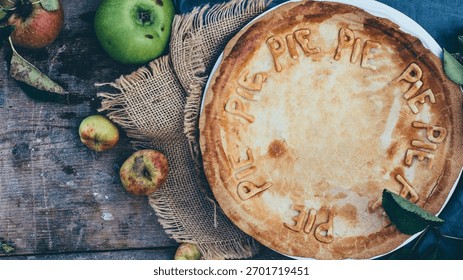 A rustic overhead view of a partially filled pumpkin pie crust surrounded by fresh apples and burlap on a weathered wooden surface.