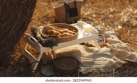 A rustic picnic setup featuring a tray of croissants, a glass of milk, and a basket of blueberries amidst a golden field of hay