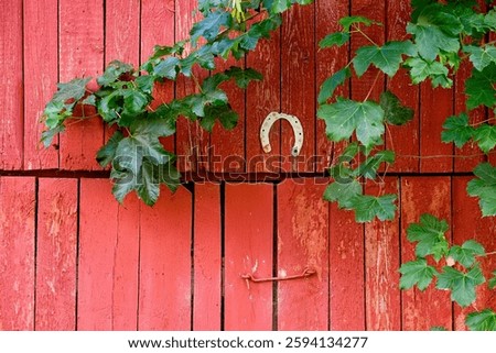 A rustic red barn door features a horseshoe and is surrounded by lush green vines on a serene countryside backdrop.