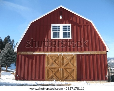A rustic red barn stands tall against the blue sky with snow on the ground around it.