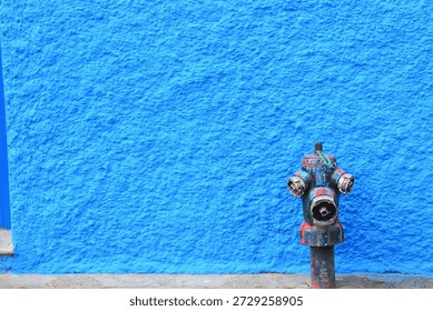 Rustic red fire hydrant with subtle rust and faded paint protrudes from a smooth vibrant blue stucco wall, creating bold color contrast in an urban setting. Dual outlets and sturdy base emphasize emergency infrastructure amid textured plaster surface. Captures street art vibe, safety symbols, or cityscape details.
