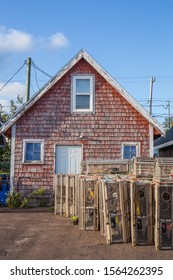 A rustic red wooden shed stands under a blue sky behind stacks of traditional lobster traps in a fishing village on Prince Edward Island, Canada.