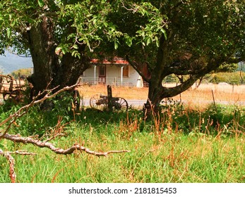 Rustic Rural Scene With Old Farm House And Wagon Wheels Framed By Large Trees And Foliage In New Zealand Countryside.