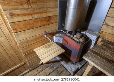 rustic sauna interior with a red brick stove loaded with stones, a metal chimney, a water tap with a red valve, and wooden benches against log walls