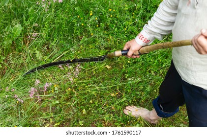 Rustic scythe on a background of green grass. The male hand holds a scythe, a sharp scythe mows the grass