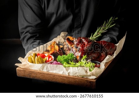 Rustic serving of roasted pork knuckle with pickled vegetables, herbs, and greens on parchment-lined tray. Traditional meat dish, ideal for restaurant, pub, or culinary stock use.
