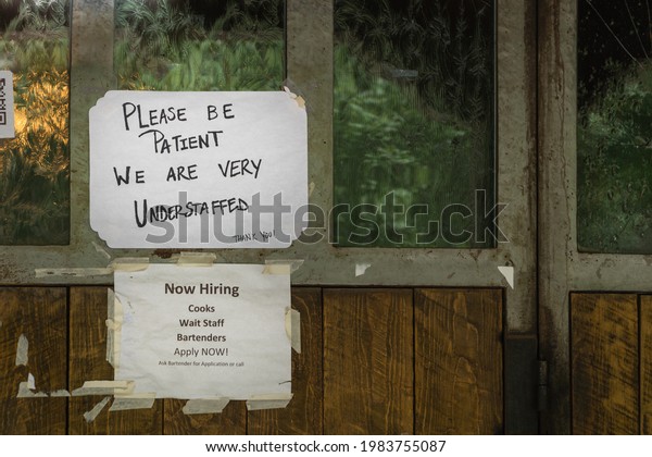Rustic sign in restaurant window during time of\
Covid stating restaurant is understaffed and is hiring in a\
struggling economy, showing the plight of small businesses in the\
post-pandemic economy.