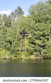 A rustic small lake cabin built on a steep forested hill featuring a long wooden staircase leading down toward a calm water shoreline.