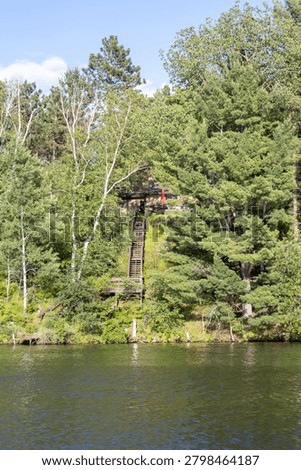 A rustic small lake cabin built on a steep forested hill featuring a long wooden staircase leading down toward a calm water shoreline.